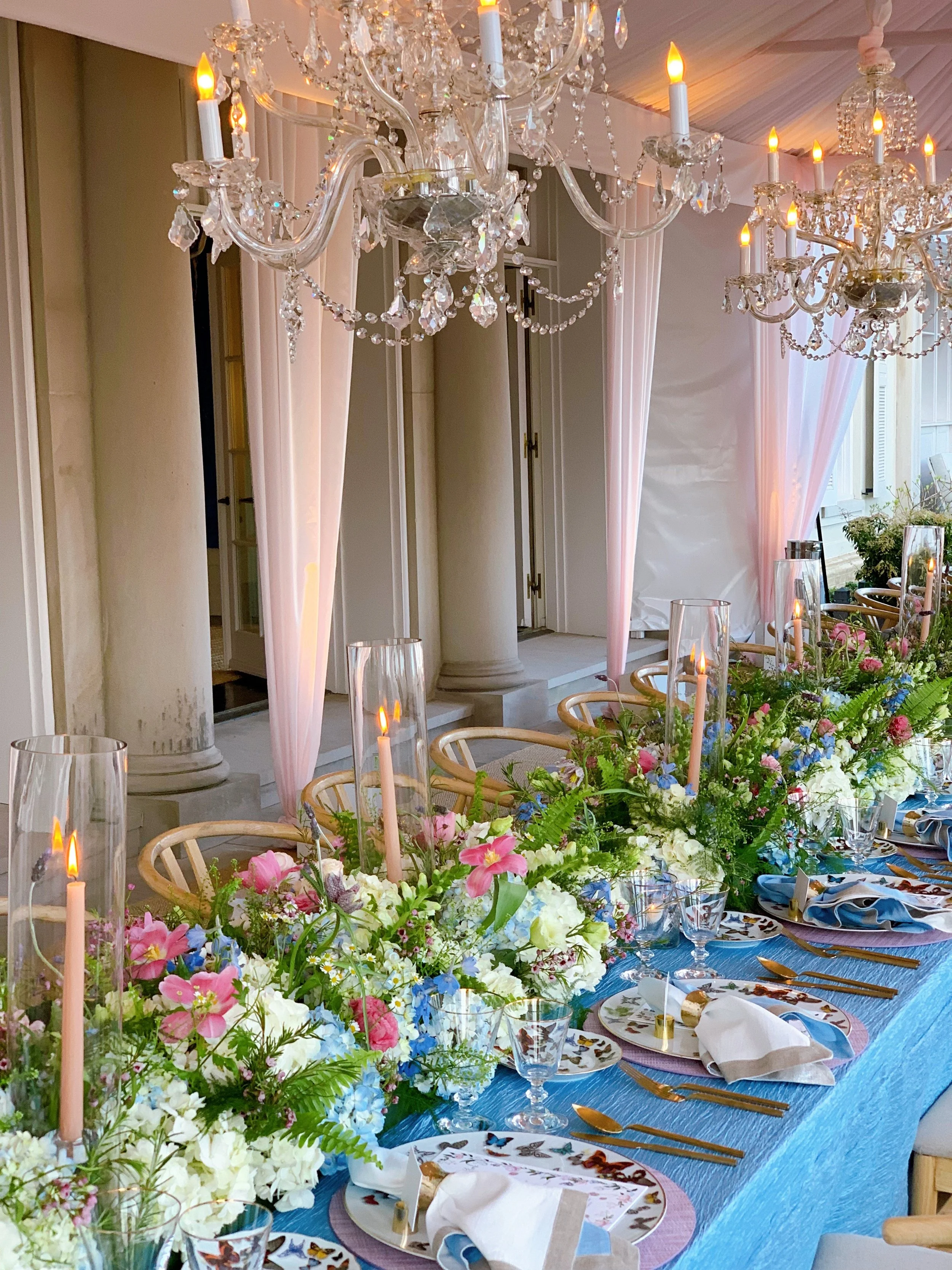 Elegant banquet table with colorful floral arrangements, lit candles, crystal chandeliers, butterfly-patterned plates, and blue table linens