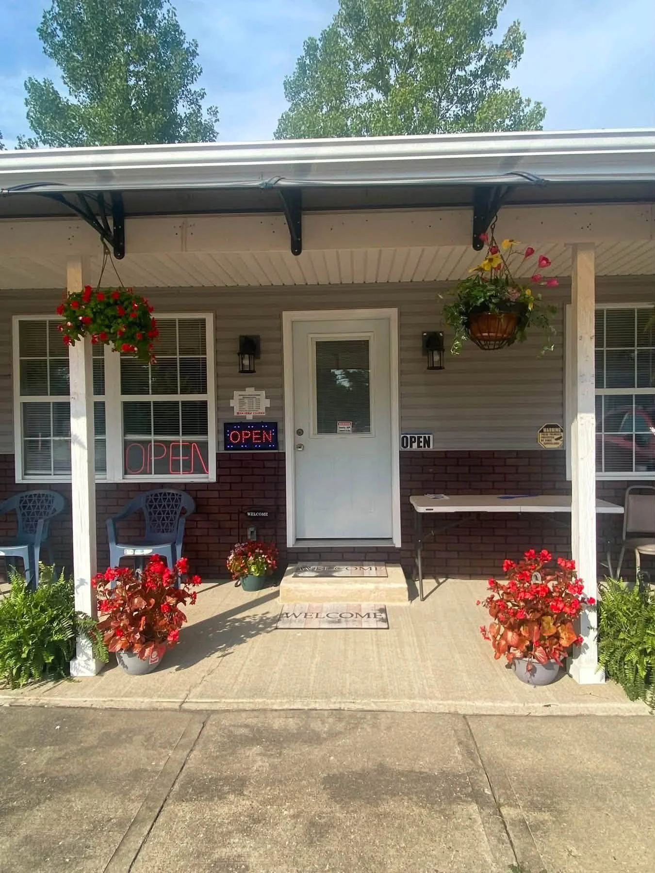 Welcoming storefront entrance with an OPEN sign, hanging flower baskets, potted plants, and outdoor seating.