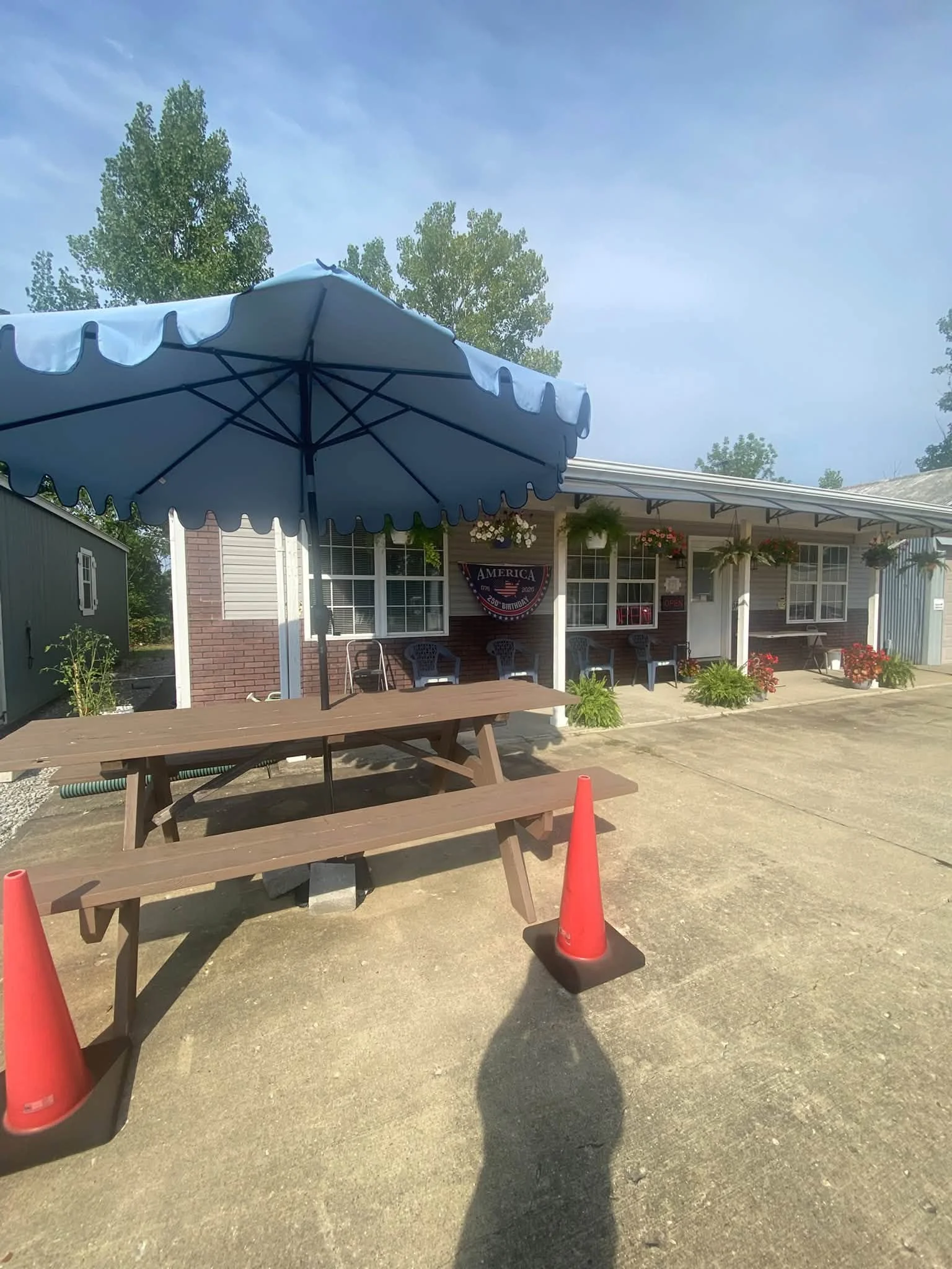 Outdoor patio with picnic tables, blue umbrella, red traffic cones, and a mobile home decorated with hanging flower baskets.