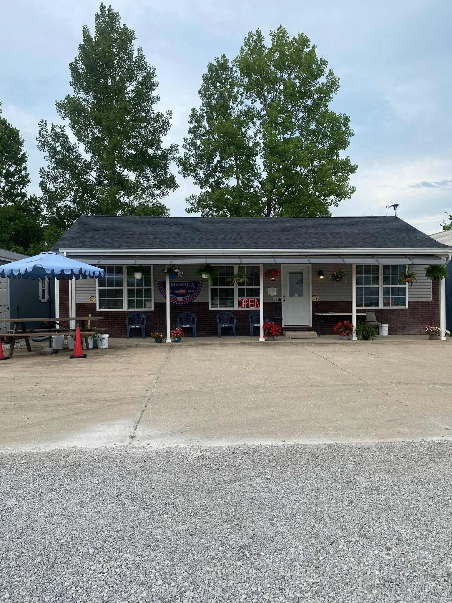 Small roadside business with an open sign, patriotic 250th birthday banner, hanging flower baskets, blue patio umbrella, and gravel parking area