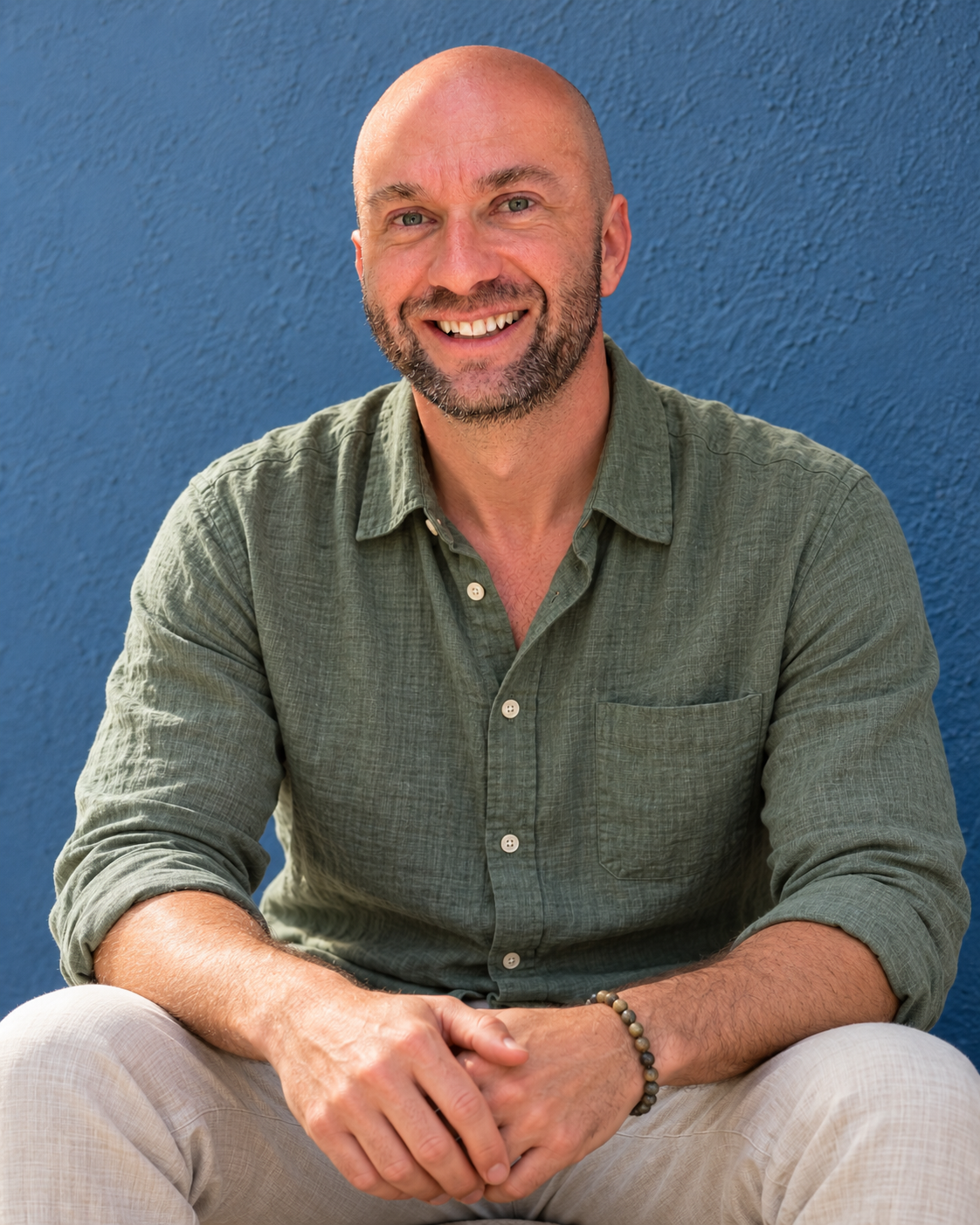 A smiling man with a beard, wearing a green button-up shirt and beige pants, sitting with his hands clasped in front of him against a blue textured wall background.