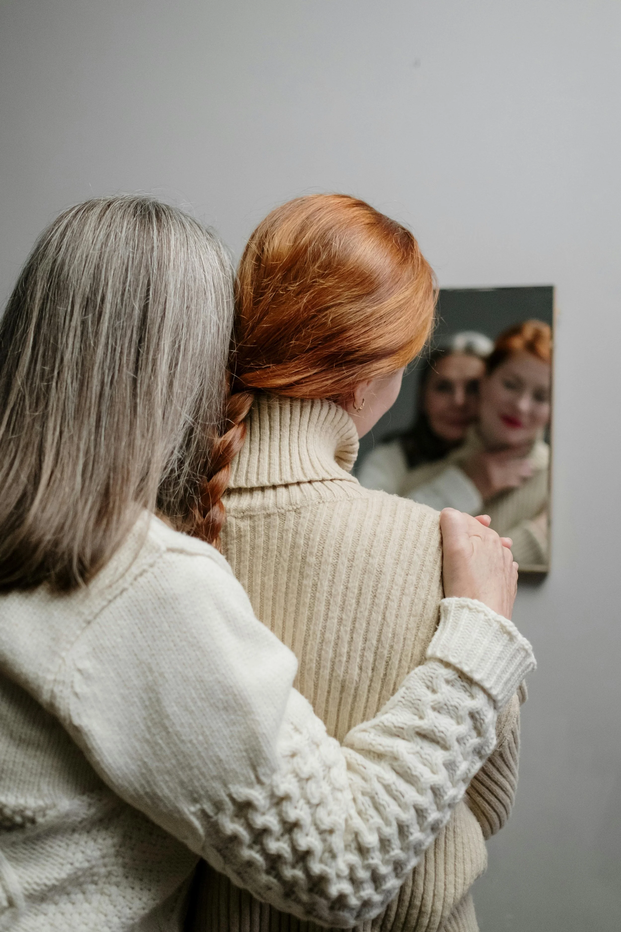 Two women in cream sweaters embrace in front of a mirror, with their reflections visible.