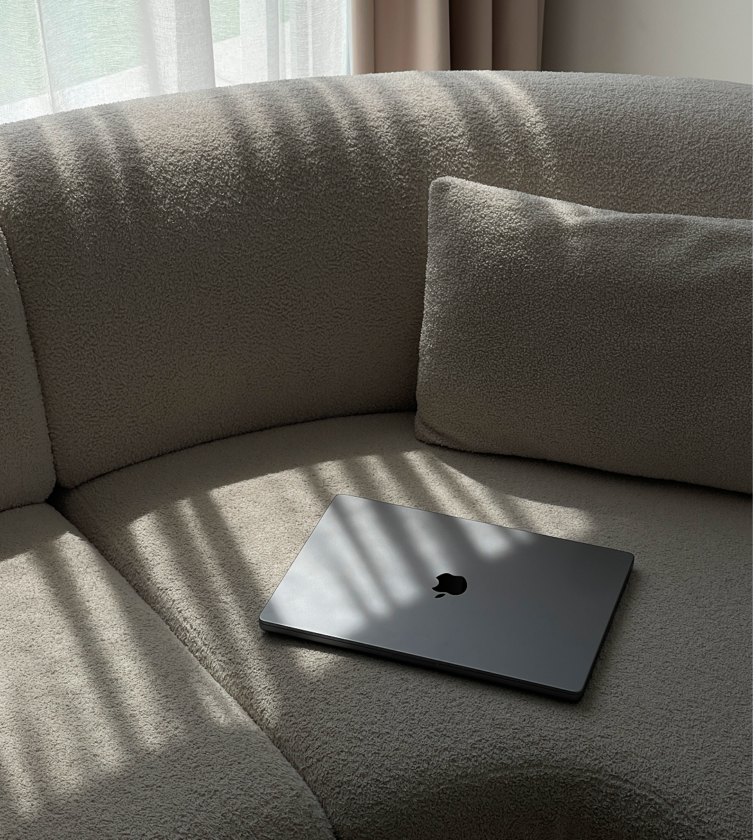 Closed MacBook on a beige textured sofa illuminated by sunlight through sheer curtains