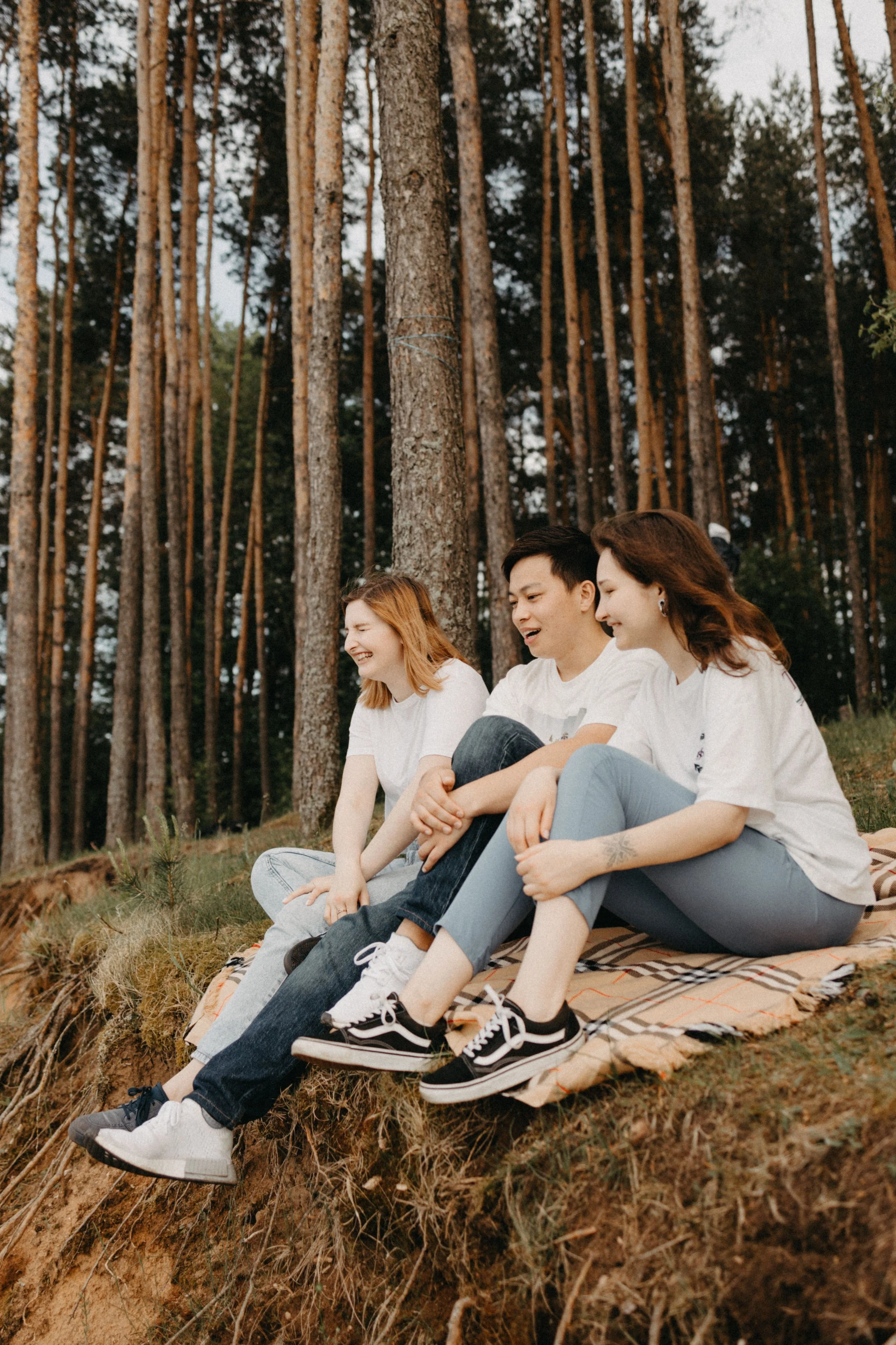 Three friends laughing together on a picnic blanket in a pine forest.