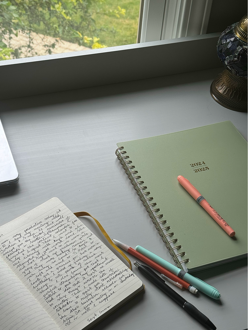 Open handwritten journal, spiral-bound 2024–2025 planner, and colorful pens on a desk beside a window
