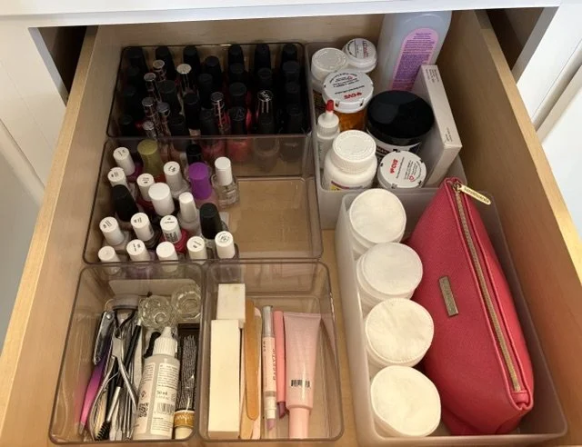 Open bathroom drawer organized with nail polish bottles, cotton pads, skincare products, makeup brushes, tweezers, and a red cosmetic pouch.