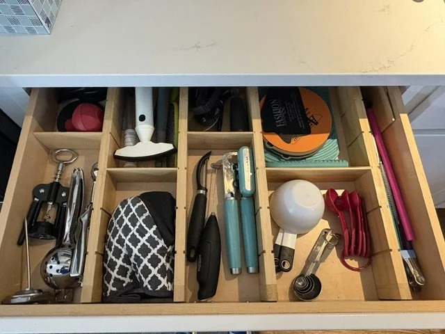 Open kitchen drawer organizer holding wine openers, utensils, measuring spoons, a whisk, and assorted household tools