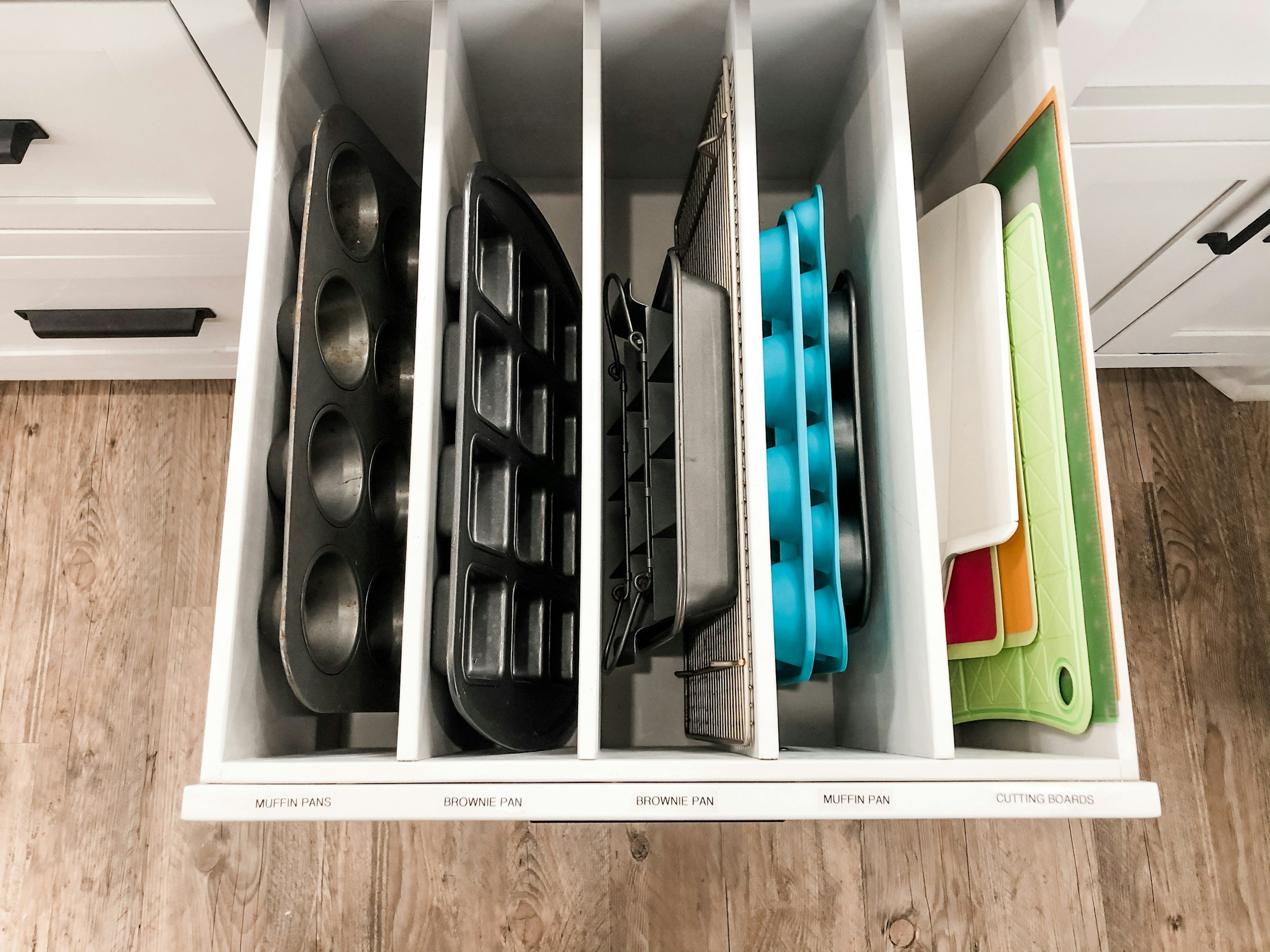 Open kitchen drawer organized with muffin pans, brownie pans, baking trays, and colorful cutting boards.