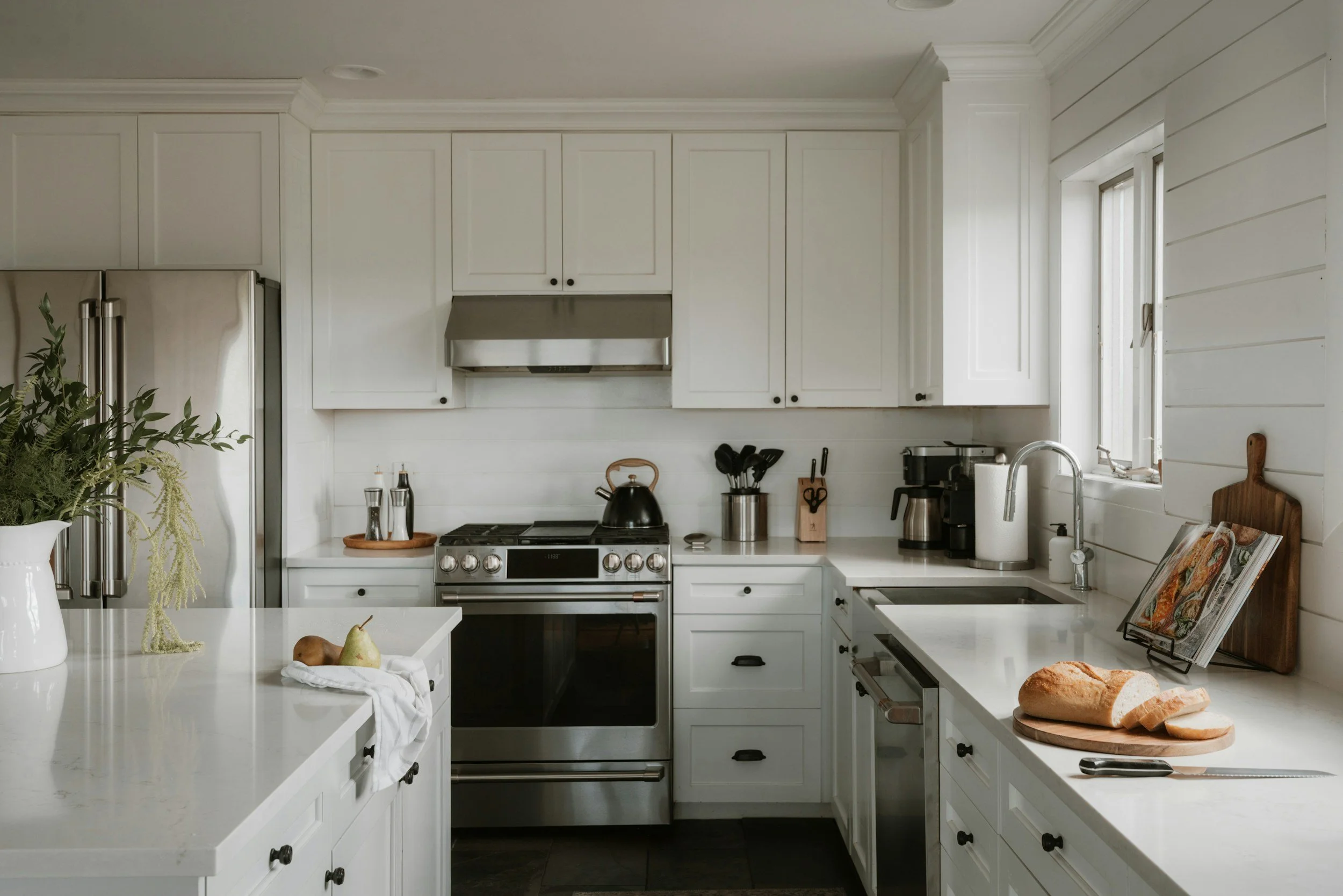Bright white kitchen with stainless steel appliances, marble countertops, farmhouse sink, cookware, fresh bread, and fruit.
