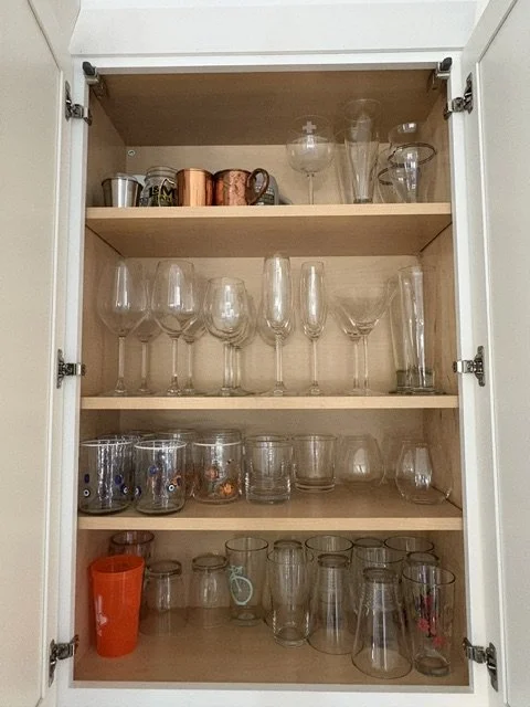 Open white cabinet displaying wine glasses, champagne flutes, tumblers, mugs, pitchers, and an orange plastic cup