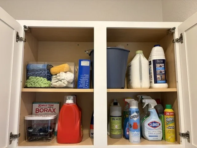 Open cabinet stocked with cleaning supplies, laundry detergent, microfiber cloths, Borax, bleach, spray cleaners, and scrub brushes