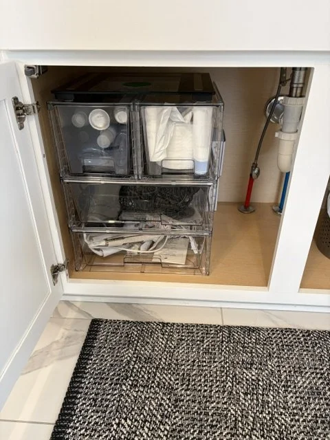 Open bathroom cabinet under the sink with clear storage bins holding cleaning supplies, toilet paper, and newspapers beside plumbing pipes.