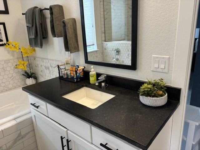 Modern bathroom vanity with black countertop, white cabinets, undermount sink, framed mirror, gray towels, and decorative plants