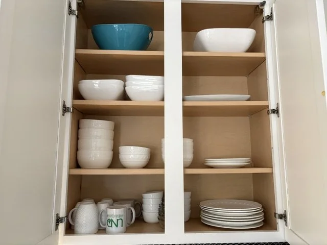 Open kitchen cabinet displaying neatly stacked white bowls, plates, mugs, and cups, plus a turquoise bowl.
