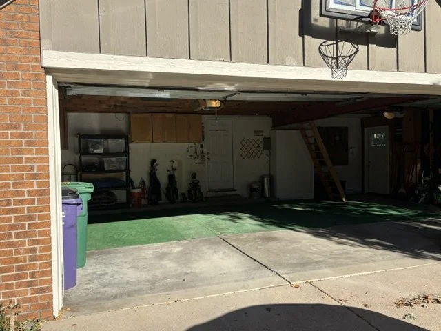 Open residential garage with a basketball hoop, storage shelves, ladder, trash bins, and green flooring