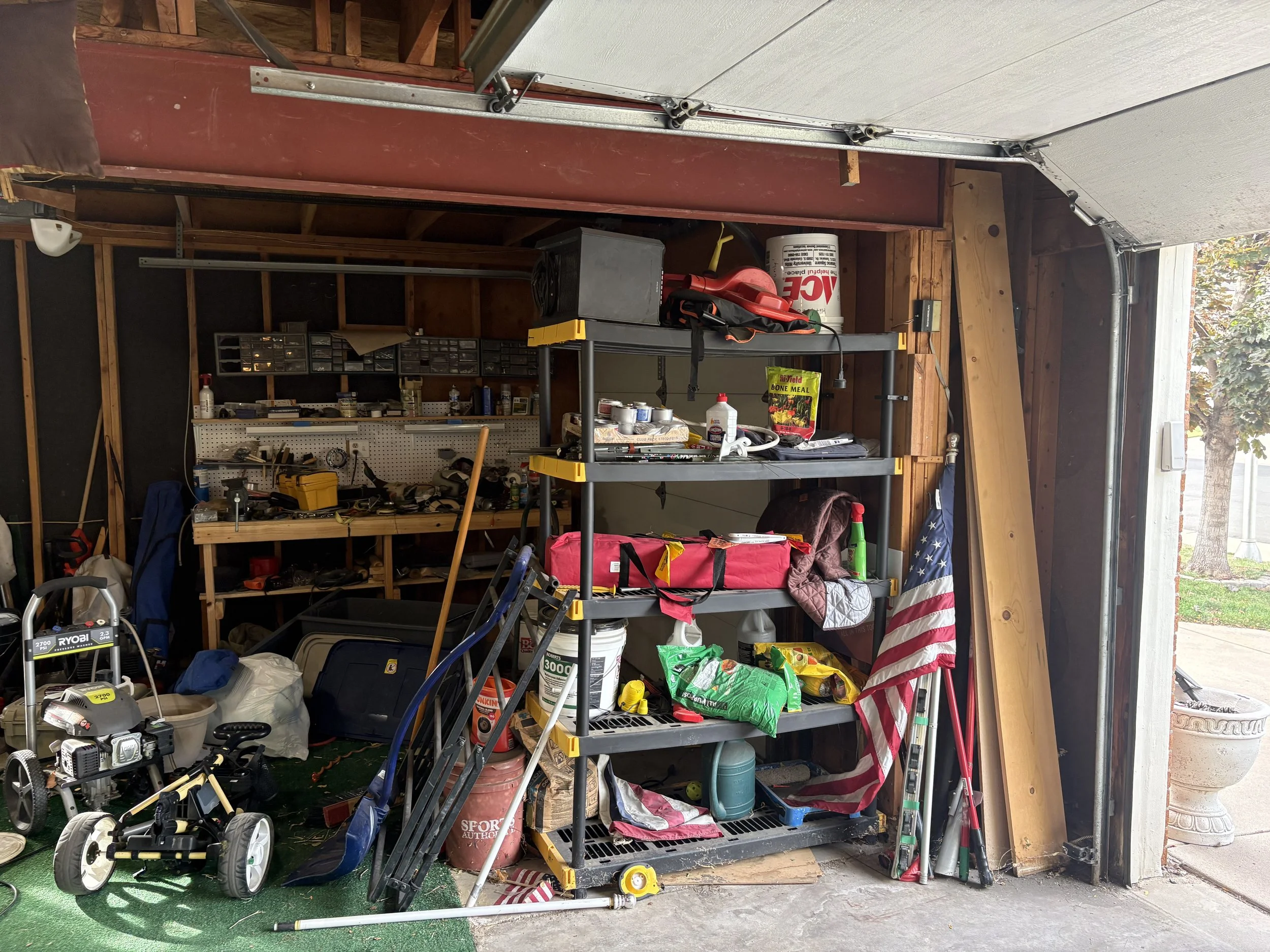Cluttered garage workshop with shelving, gardening tools, lawn equipment, storage bins, and an open garage door