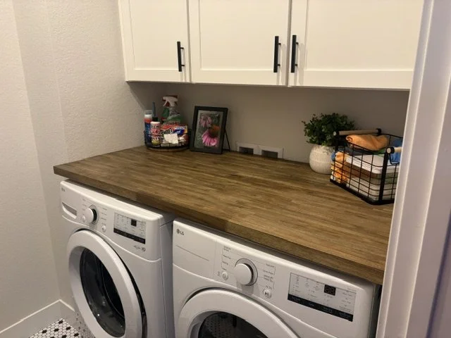 Compact laundry room with a front-loading washer and dryer beneath a wood countertop, white cabinets, cleaning supplies, framed artwork, and a potted plant