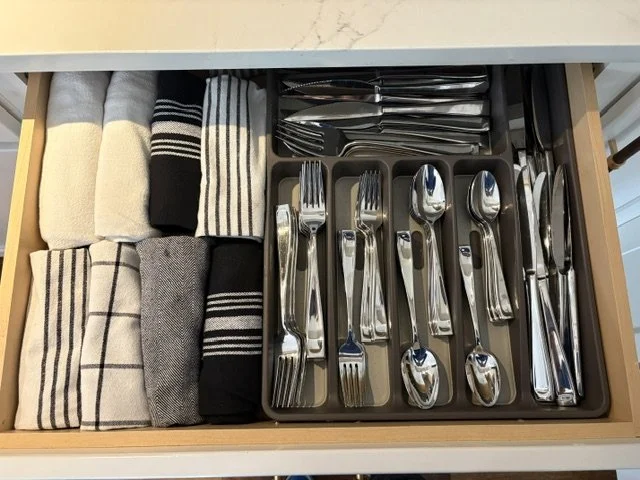 Open kitchen drawer containing neatly folded dish towels and organized stainless steel flatware in a utensil tray
