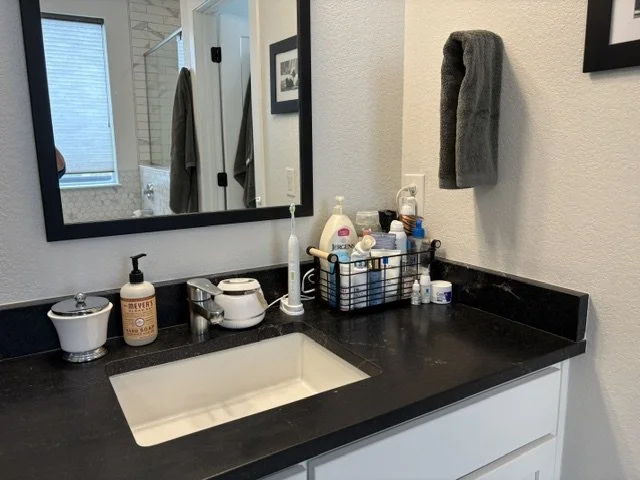 Modern bathroom vanity with undermount sink, black countertop, large mirror, gray towel, soap dispenser, and toiletries basket