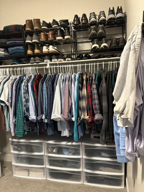 Organized walk-in closet with rows of button-down shirts, shoes on shelving, folded clothes, and storage drawers.