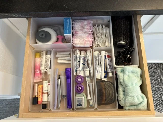 Organized bathroom drawer containing cosmetics, skincare products, cotton swabs, hairbrushes, hair ties, and washcloths