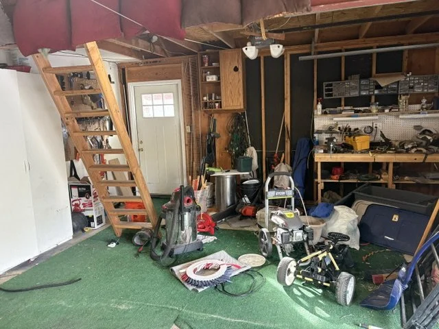 Cluttered garage workshop with lawn equipment, tools, shelving, workbench, ladder, and green floor mat