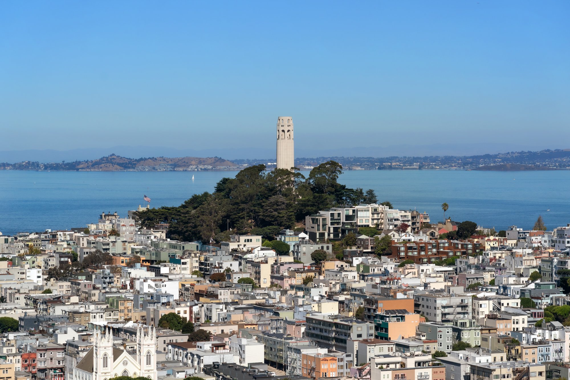 Cityscape view showing a tall tower in the distance, a large body of water, and a dense area of residential buildings and houses in the foreground with a church structure visible at the bottom left.