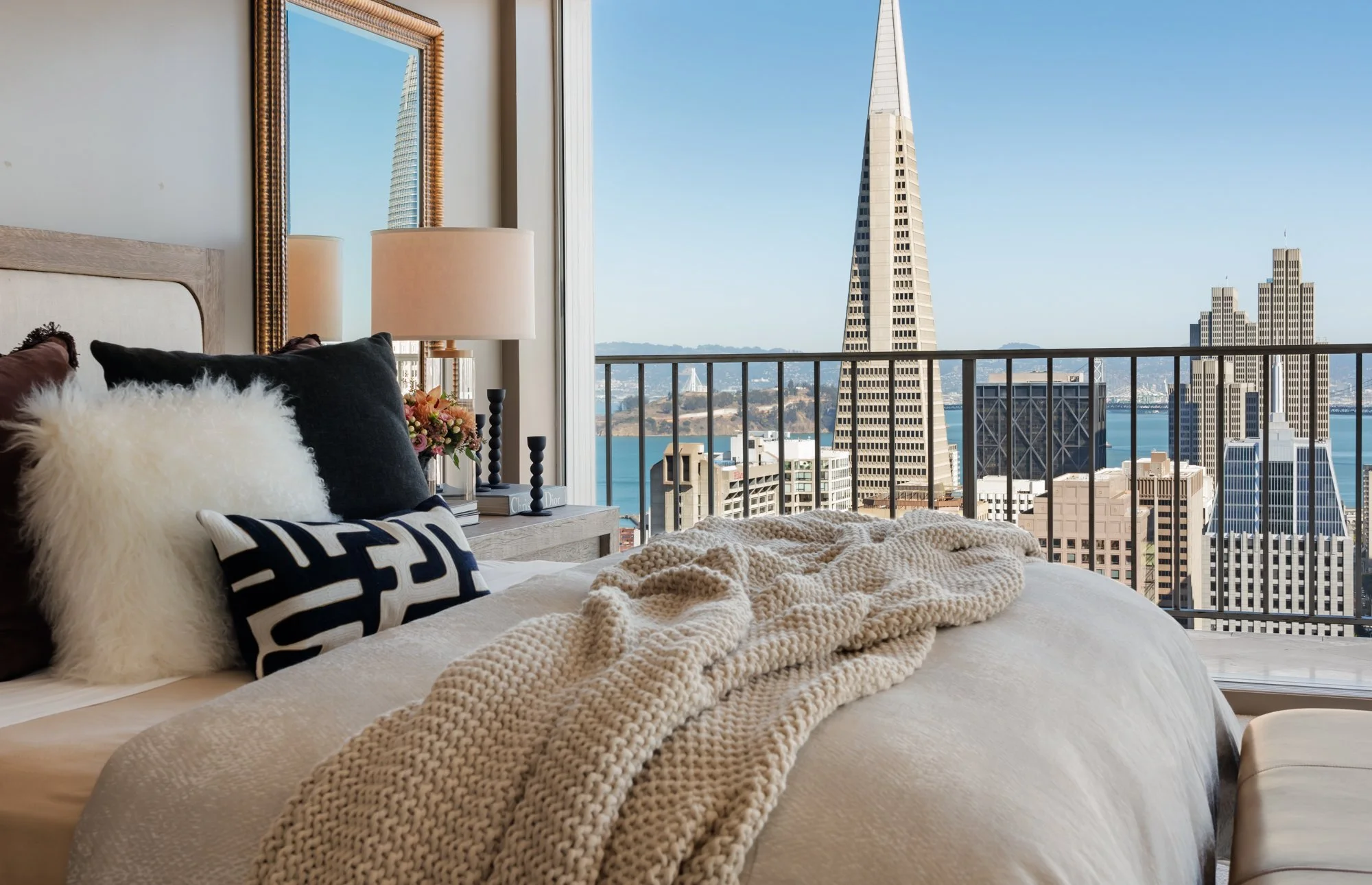 A bedroom with a bed featuring various pillows and a textured blanket, next to a large window with city and water views, including the Transamerica Pyramid building in San Francisco.