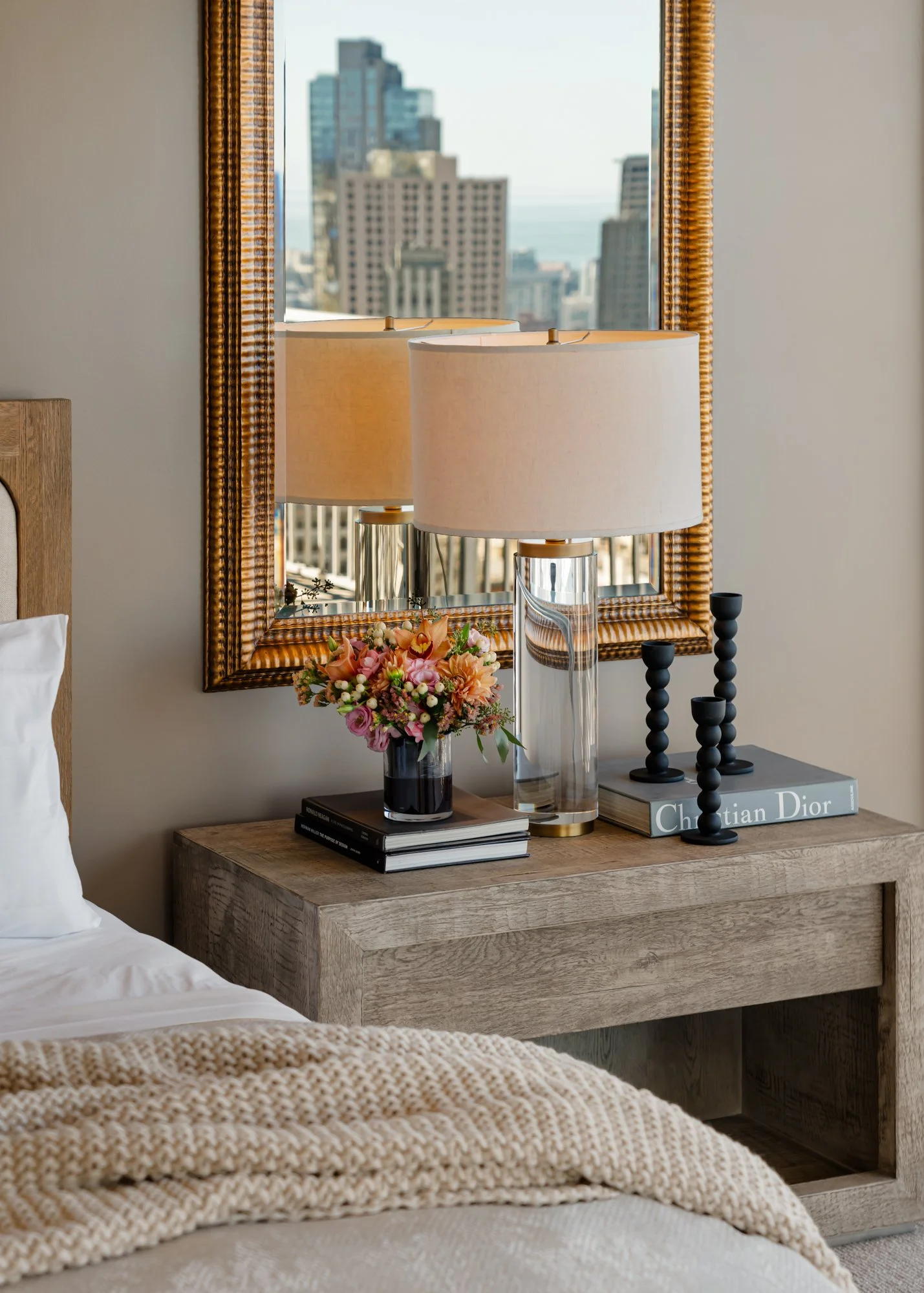 A bedside table with a flower arrangement, a glass lamp, and two black candle holders, with a mirror reflecting city buildings and a window in the background.