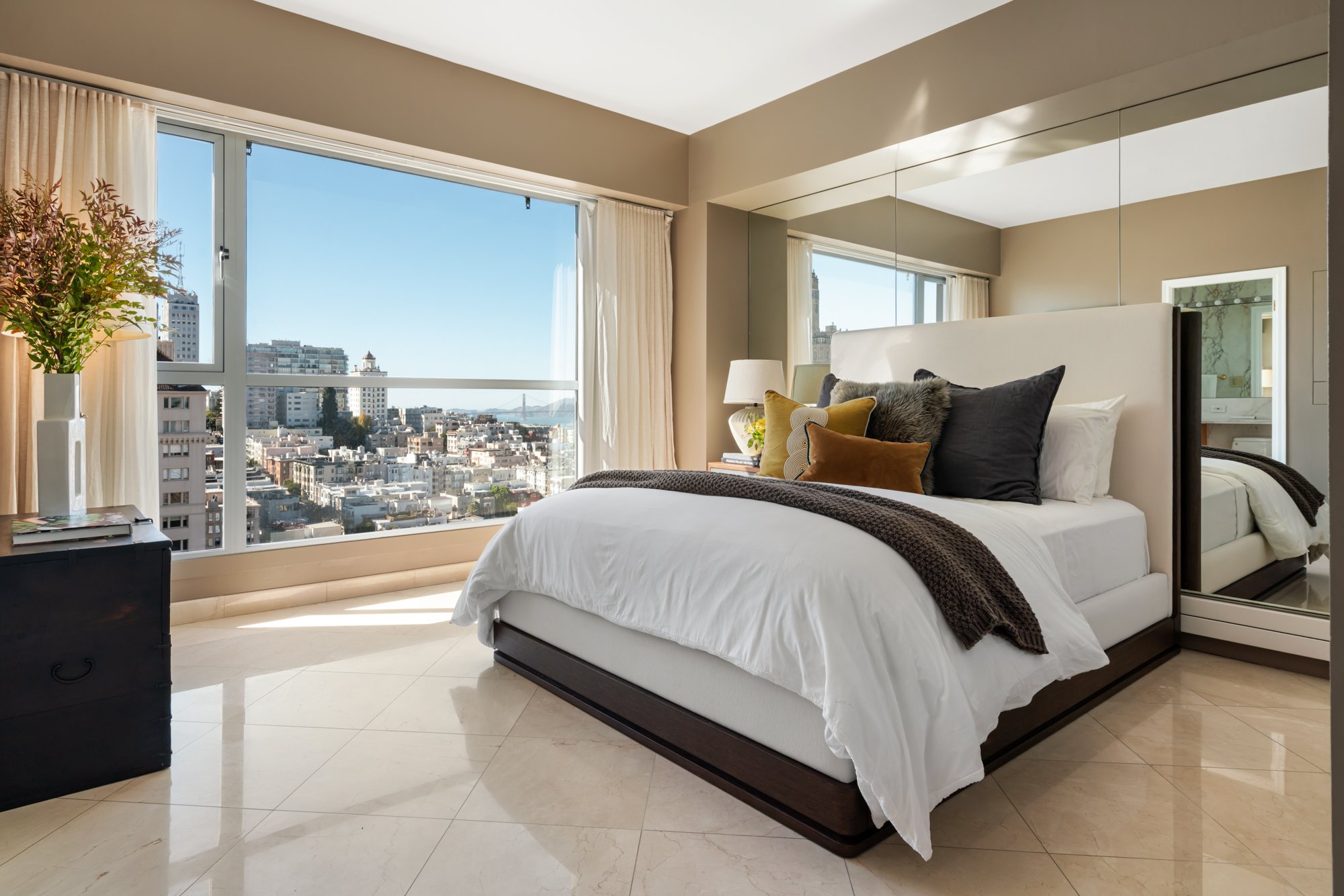 Modern bedroom with a large window overlooking city buildings and the San Francisco Bay, featuring a white bed with multiple pillows, a side table with a lamp, a potted plant, and a mirrored closet door.