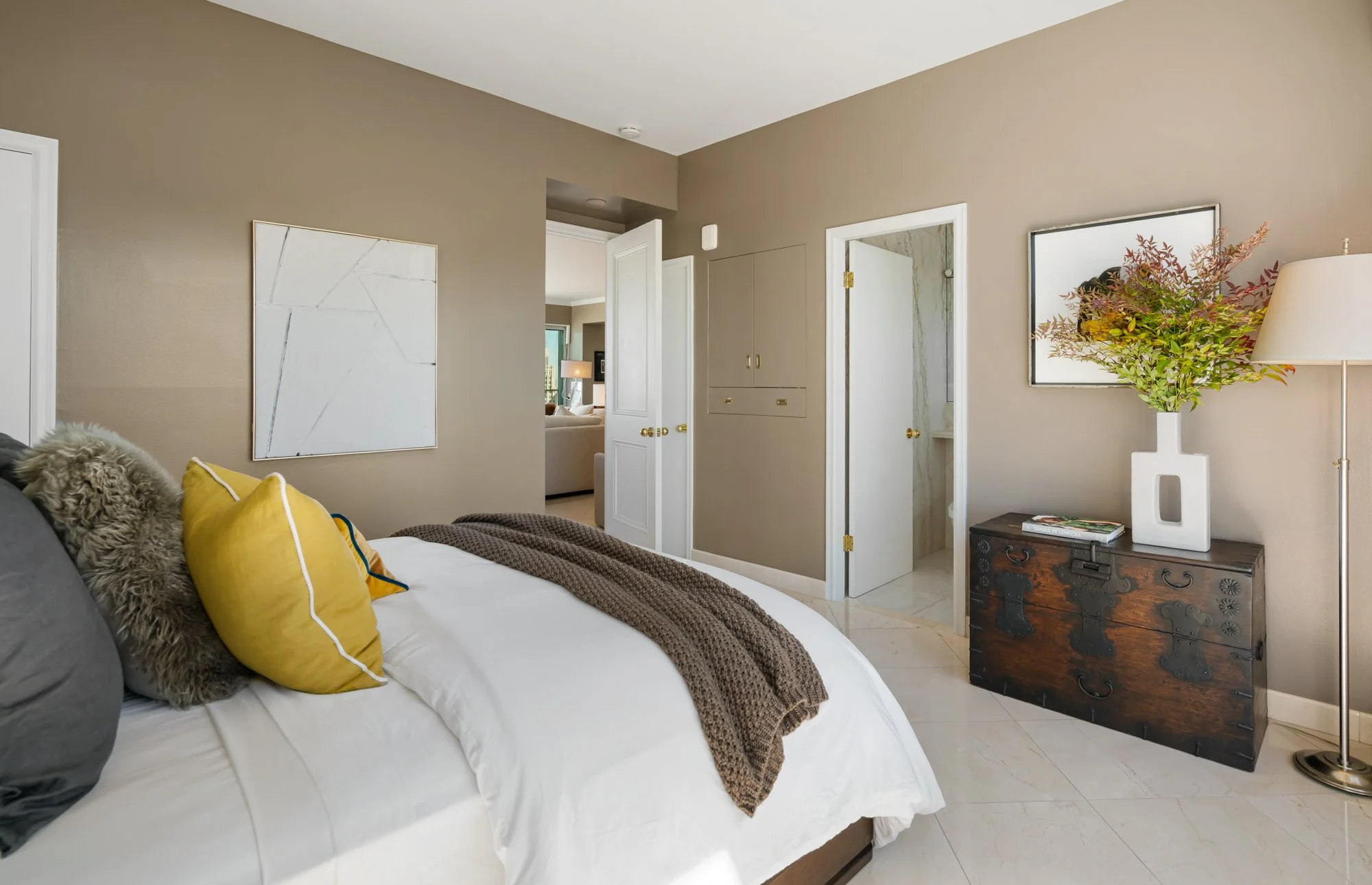 A bedroom with beige walls, white trim, and light-colored floor tiles. A bed with white bedding, yellow pillows, and a brown throw blanket is on the left. There are abstract artwork and a large mirror on the walls. A wooden chest with metal accents, 