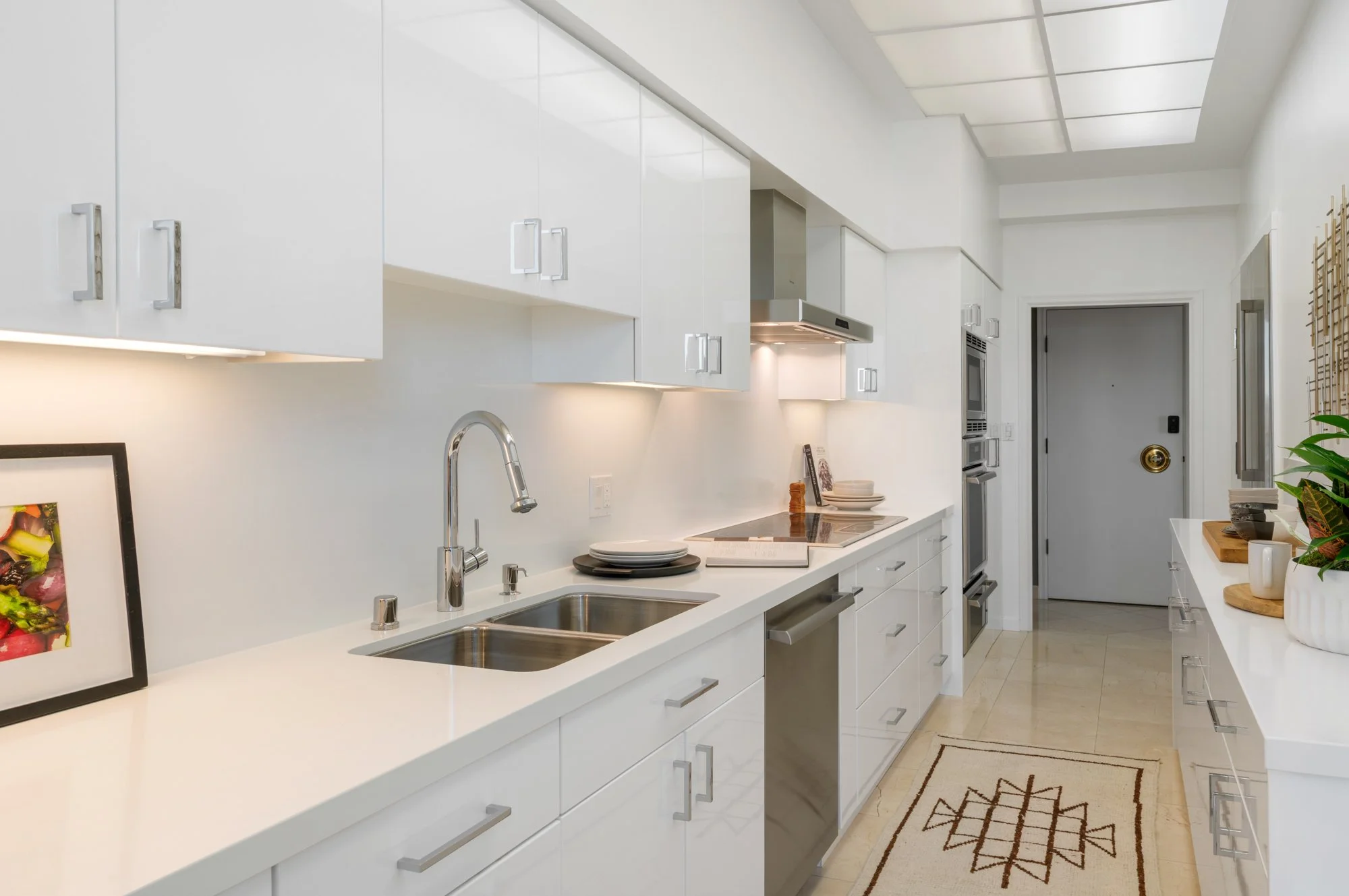 Modern white kitchen with white cabinets, stainless steel appliances, and a marble countertop with stacked plates, a sink, and decorative plants.