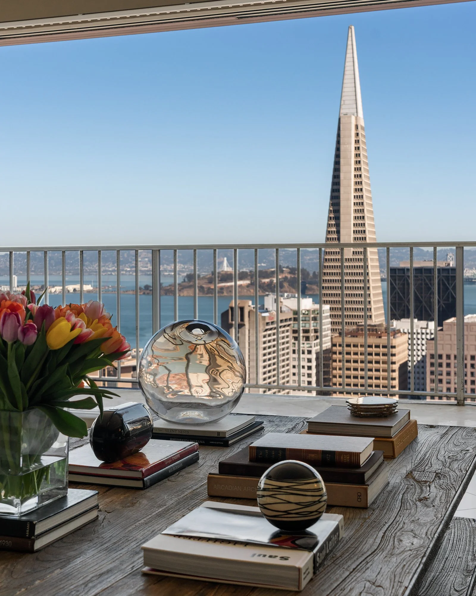 A view of a city skyline through a balcony railing, with a table in the foreground holding books, decorative objects, and a vase of colorful tulips.