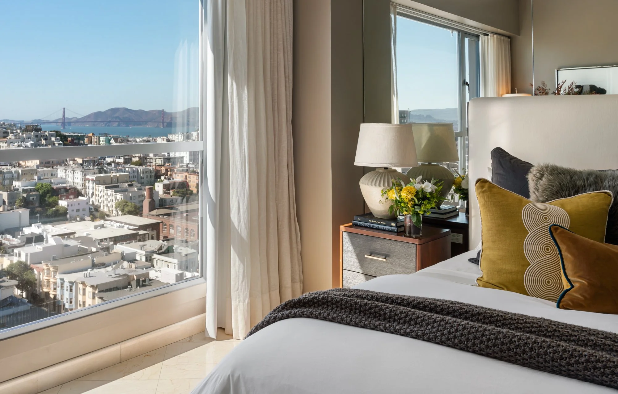 A hotel room with a large window overlooking a cityscape and a bridge in the distance. The room has a bed with multiple cushions, a bedside table with a flower arrangement, lamps, and books. Curtains are partially open, allowing sunlight to brighten 