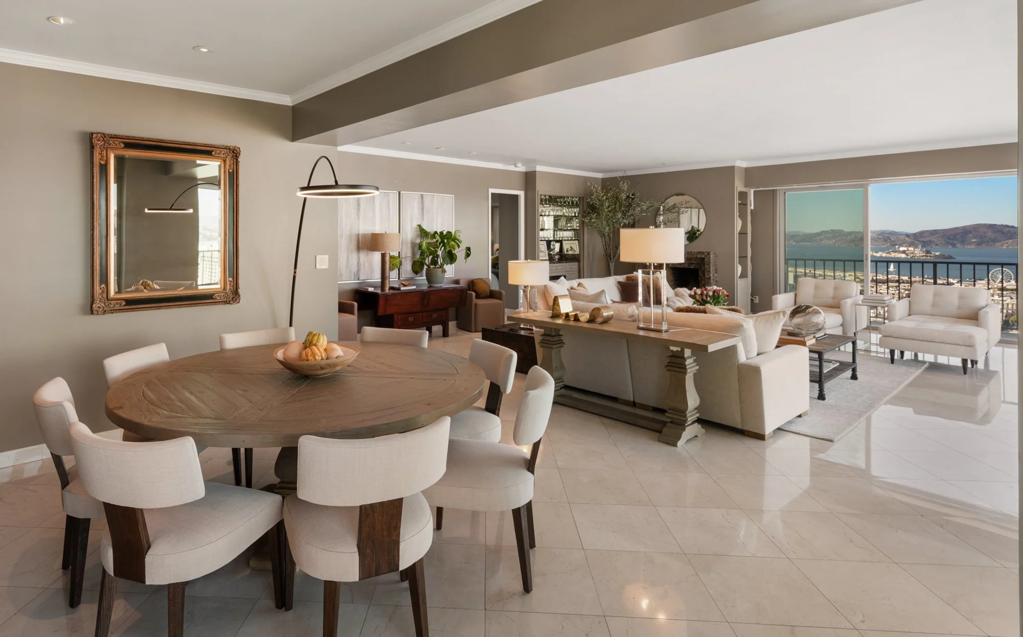Open-plan living and dining area with a round wooden dining table, beige chairs, a beige sofa, white armchairs, and large windows with a view of water and mountains.