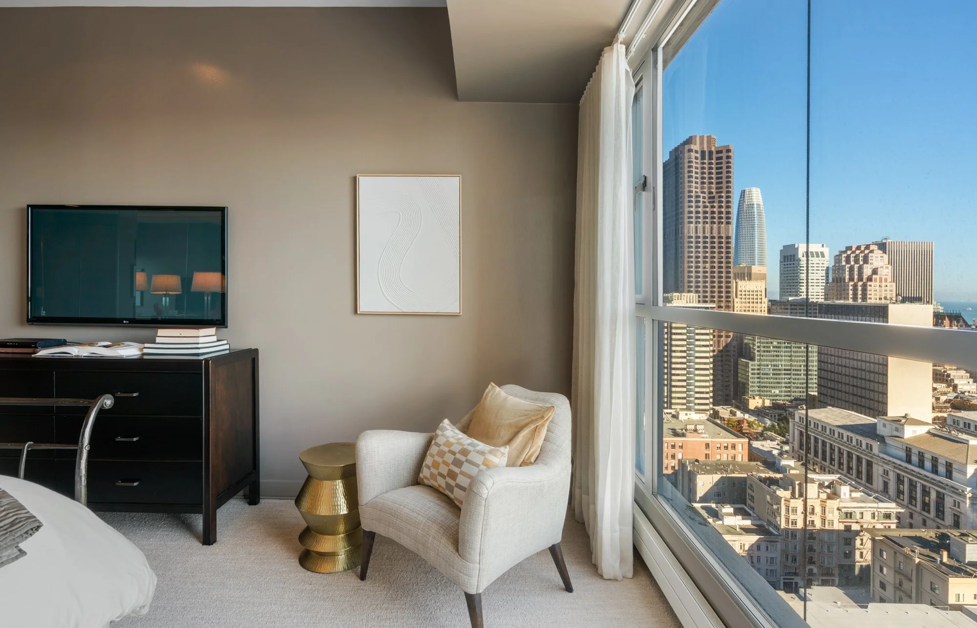 A corner of a modern living room with large windows showing a cityscape with tall buildings. The room has a beige armchair with patterned pillows, a gold side table, and a black dresser with books and a flat-screen TV mounted on the wall.