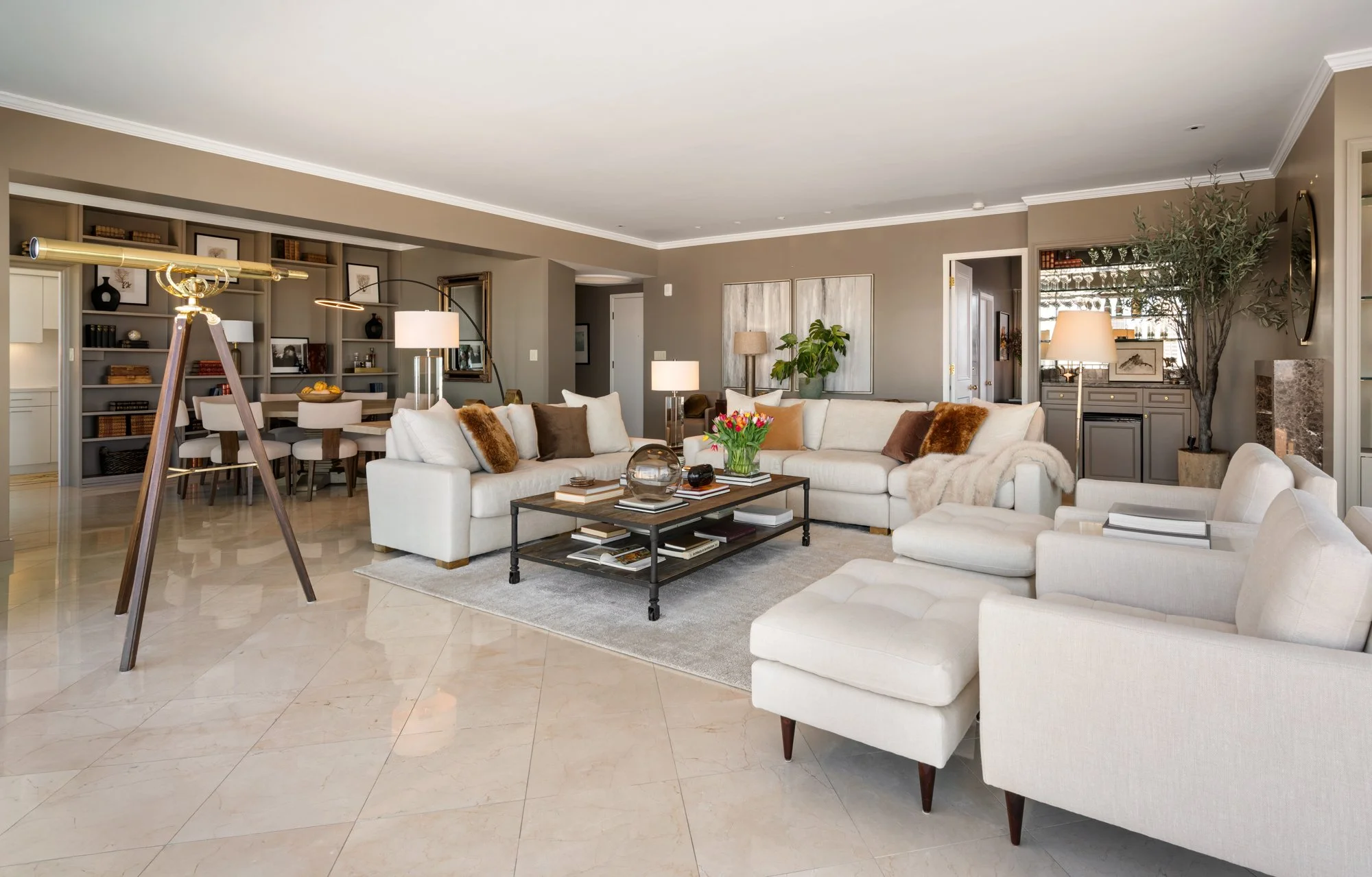 Spacious living room with beige walls, white ceiling, large white sectional sofa, glass coffee table with books and a flower vase, multiple table lamps, a large potted plant, built-in bookshelves, and a dining area in the background.