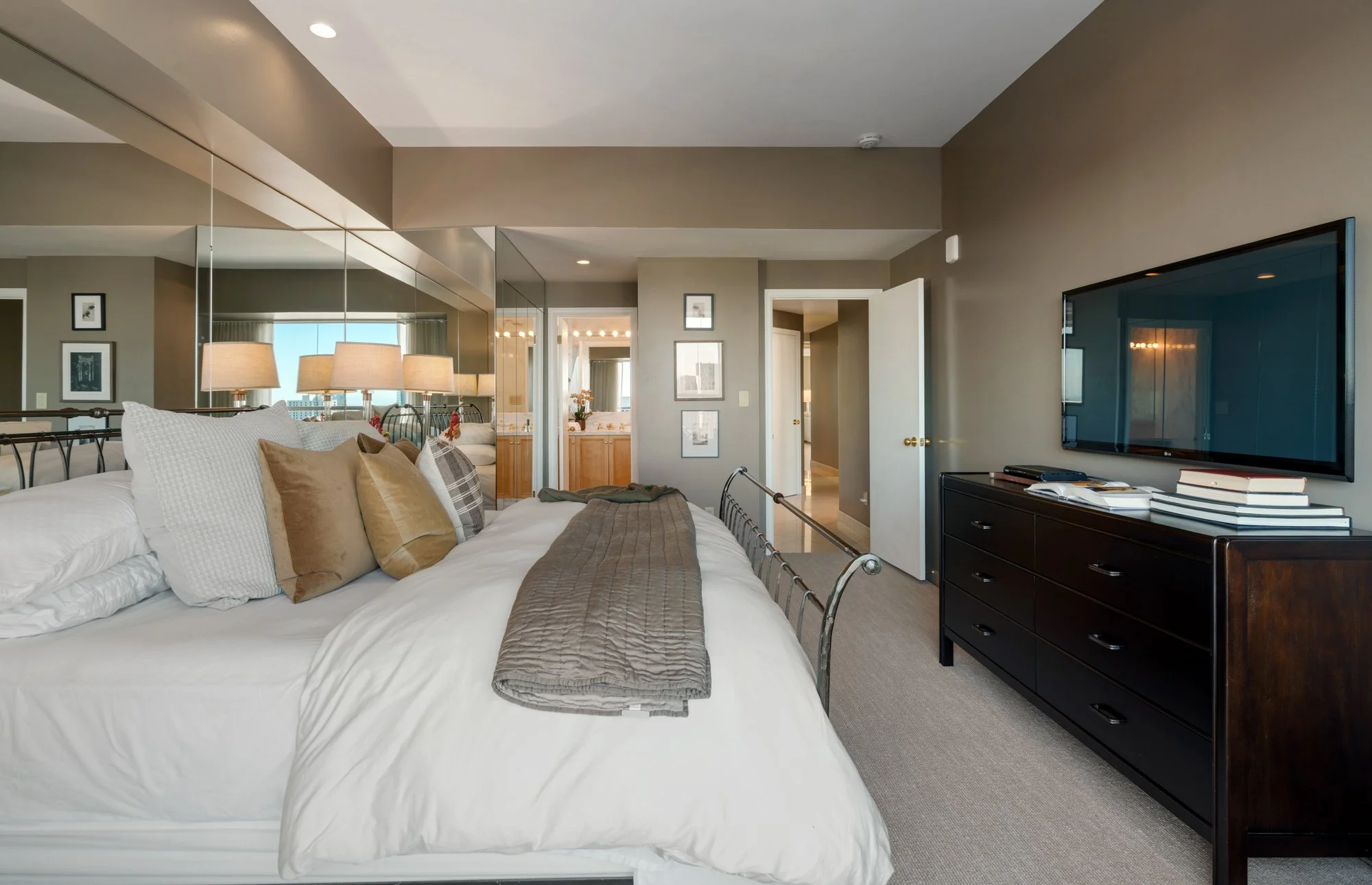 A bedroom with a large bed covered in white linens and beige and plaid pillows, a dark wooden dresser with books and a TV, and a mirrored wall reflecting the room. The walls are painted beige, and the room has a doorway leading to a bathroom or close