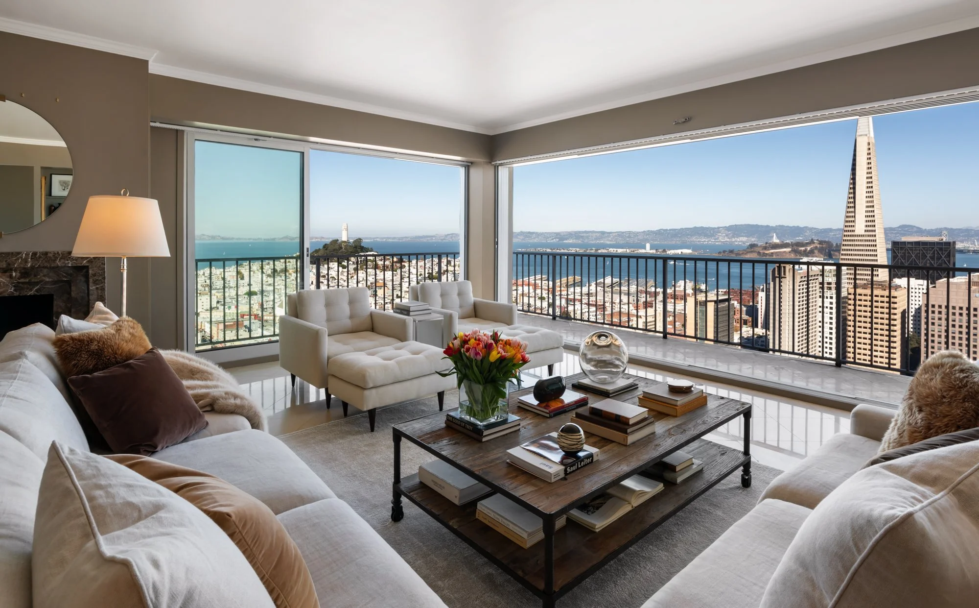 Living room with large windows showing a cityscape view including the Golden Gate Bridge and San Francisco skyline, furnished with white sofas, a wooden coffee table with books and decorative objects, a lamp, and a vase of tulips.