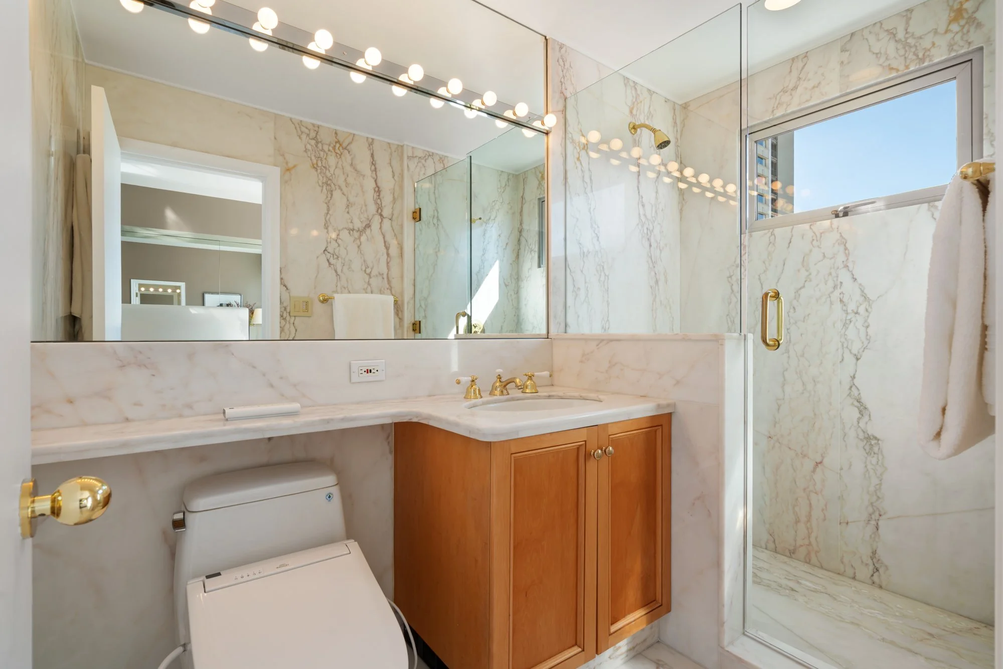 Bathroom with marble walls, a large mirror, a marble vanity with a gold faucet, a window with a city view, and a glass shower enclosure with a gold handle.