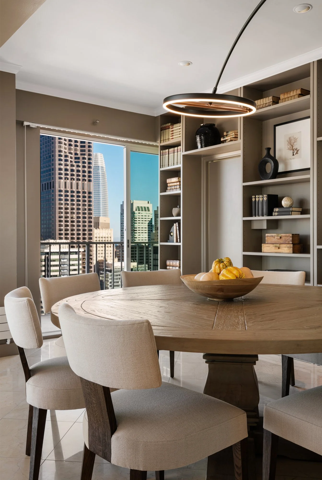 Interior view of a dining room with a round wooden table surrounded by six beige upholstered chairs, a bowl of decorative pumpkins on the table, large window with a cityscape view, and a modern black circular ceiling pendant light.