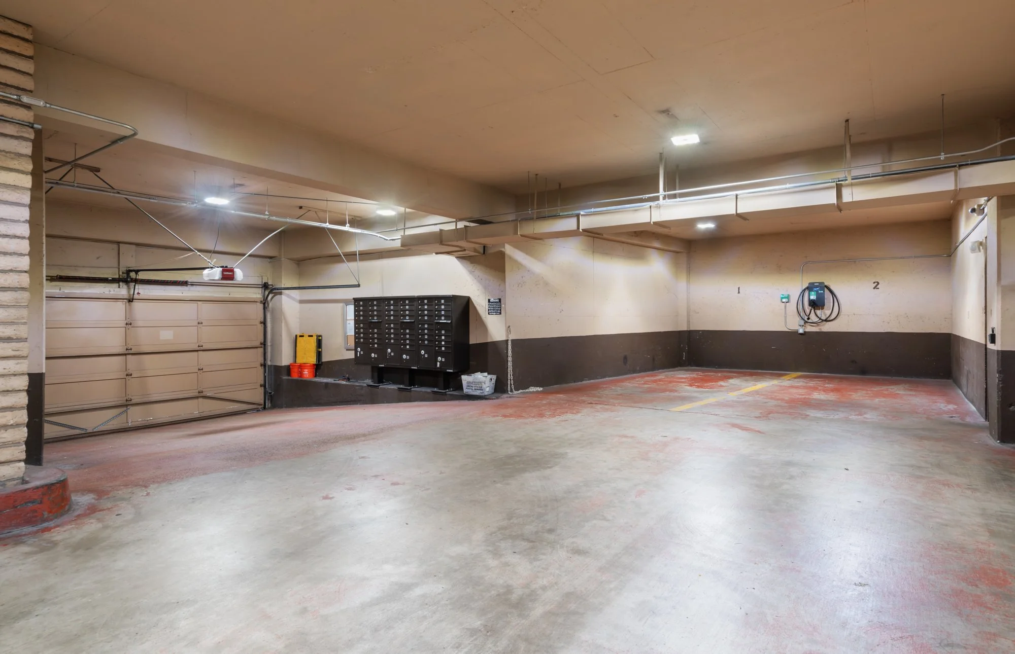 Underground parking garage with a closed garage door, mailboxes, parking spaces, electrical conduit, and lighting fixtures