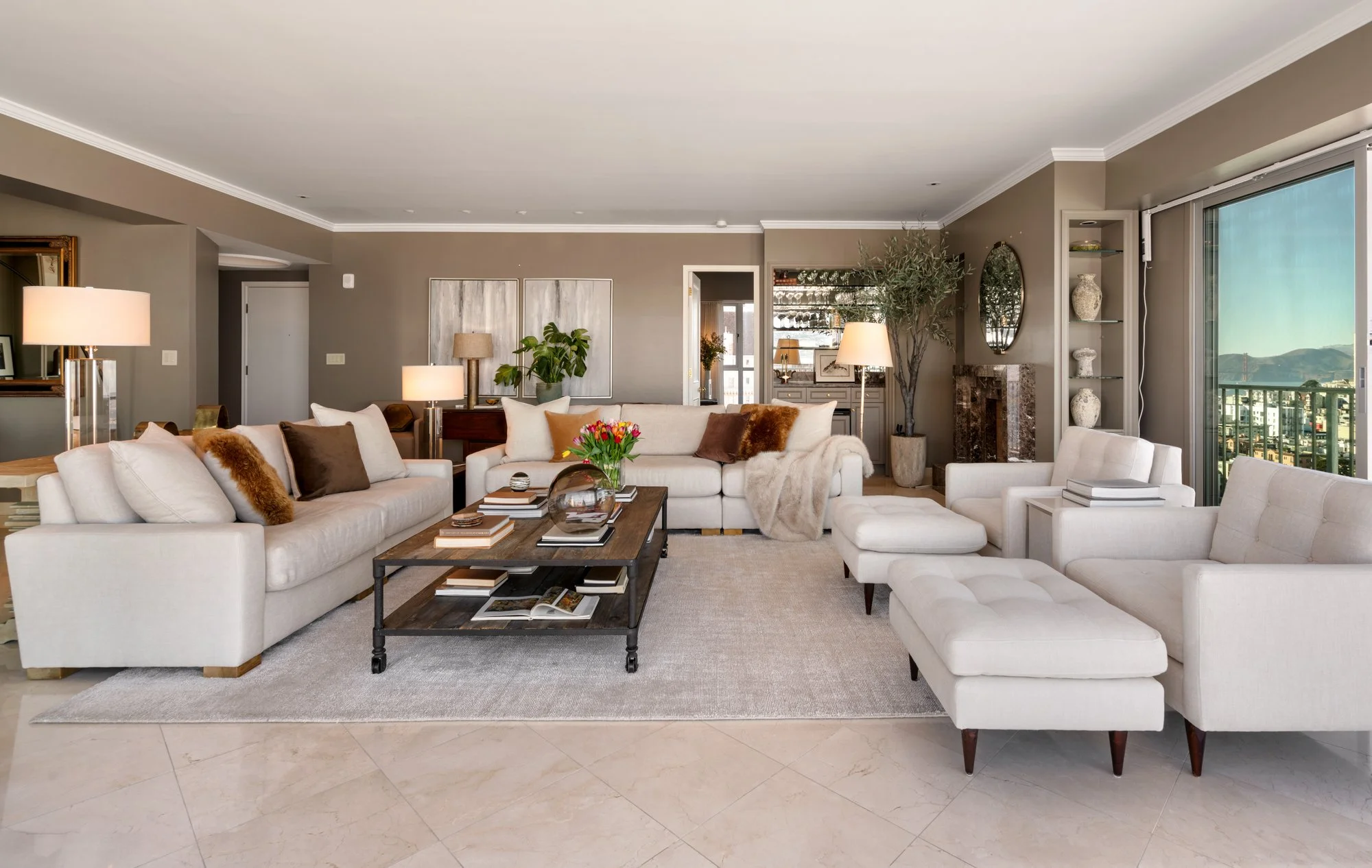 Living room with white sofas, a coffee table with books, and a large window showcasing city and mountain views.