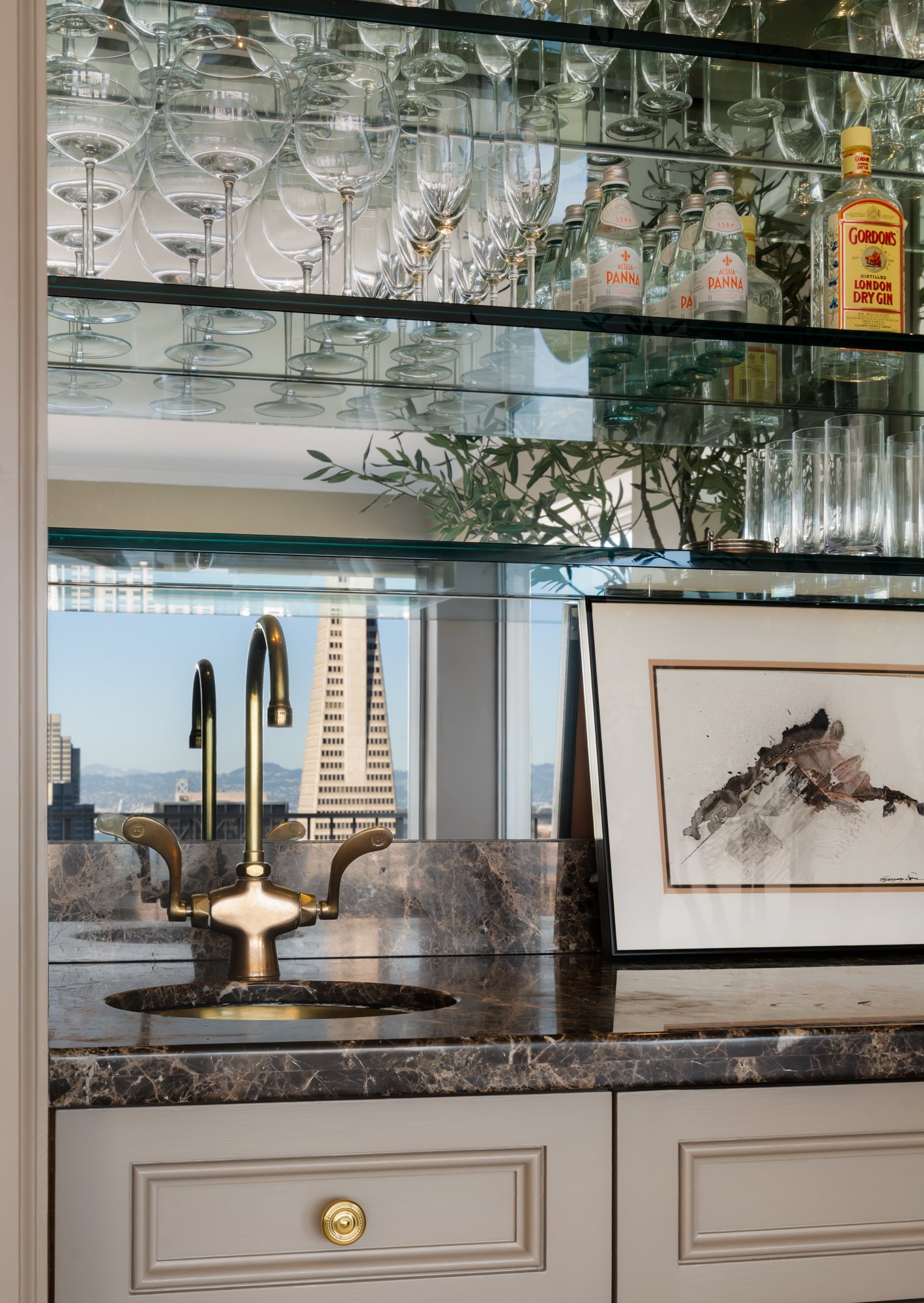A modern kitchen with glass shelves holding wine glasses, champagne flutes, and bottles of Acqua Panna water and Gordon's London Dry Gin. A framed artwork is placed on the countertop, and a cityscape with tall buildings and mountains is visible throu