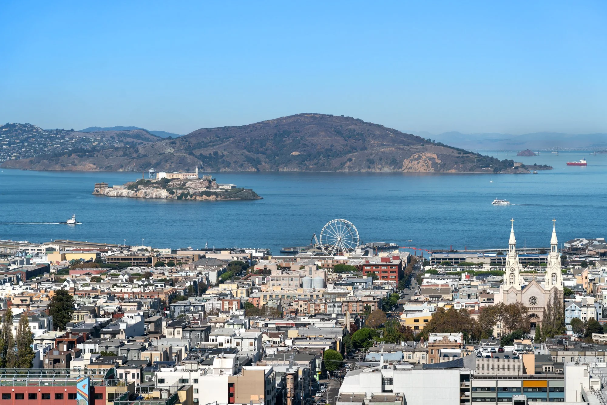 A panoramic city view with a body of water in the background, including boats, hills, and a bridge, featuring buildings, a ferris wheel, and churches with twin spires in the foreground.