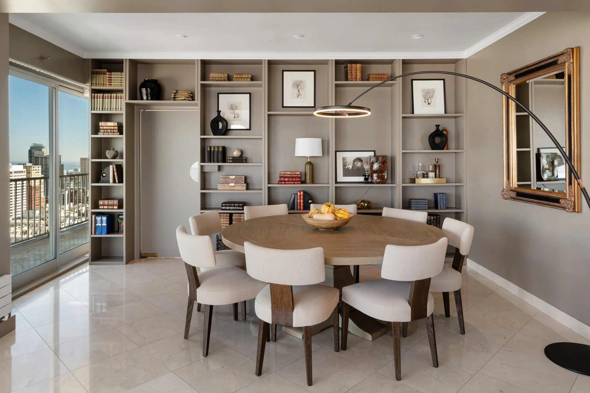 A dining room with a round wooden table surrounded by seven beige upholstered chairs, a large bookshelf with decor and books, a modern arc floor lamp, framed artwork on the wall, a white table lamp, and a balcony with a city view.