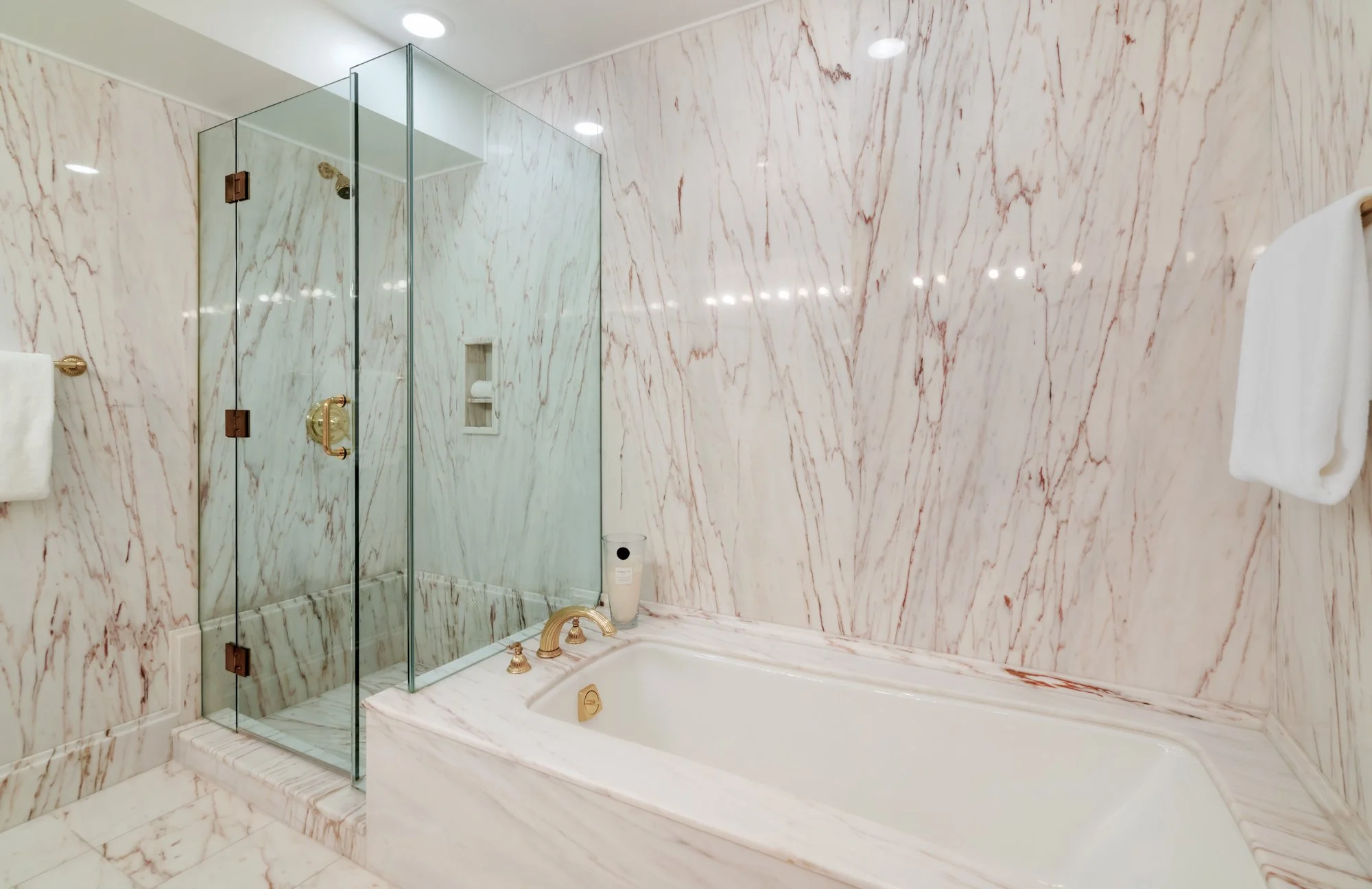 Bathroom with pink and white marble walls, a glass shower enclosure with gold hardware, and a bathtub with a matching marble surround.