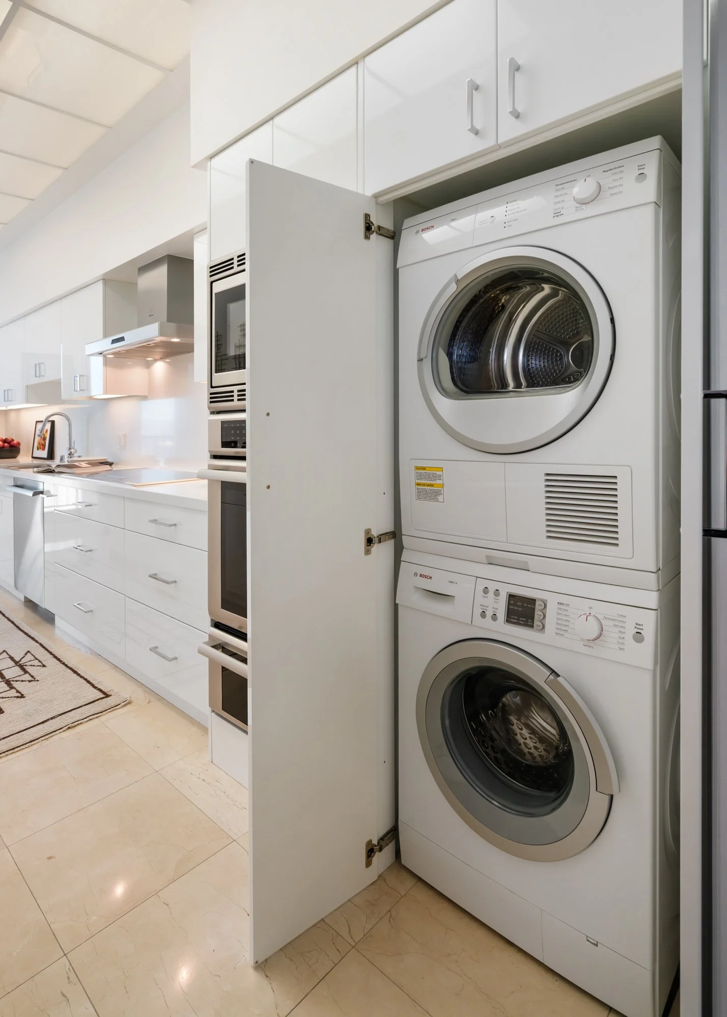 Stacked washer and dryer in a kitchen closet with white cabinets and beige tiled floor.