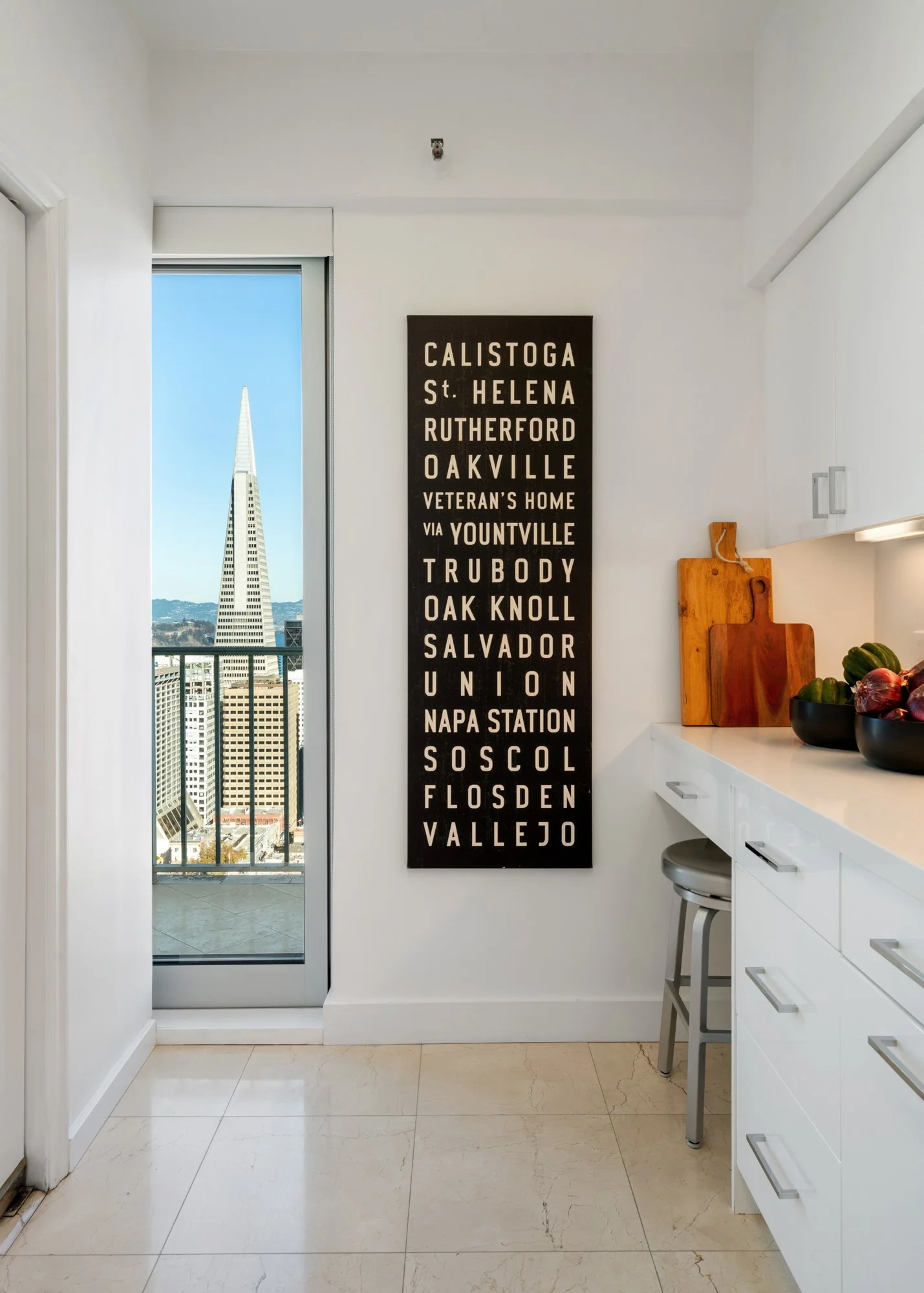 Interior view of a modern kitchen with white cabinets, a marble countertop, and a view of the San Francisco skyline with the Transamerica Pyramid through a sliding glass door. A black wall art piece lists San Francisco neighborhoods and landmarks in 