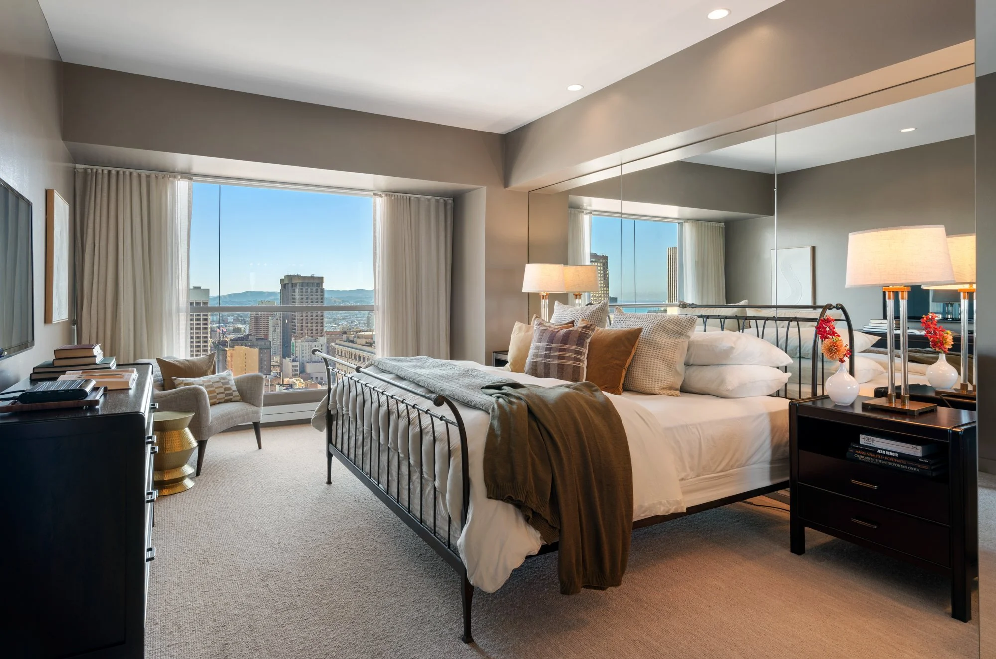 A bedroom with a metal bed frame, cream bedding, and pillows, illuminated by natural light from large windows showing a city view with tall buildings and mountains in the distance.